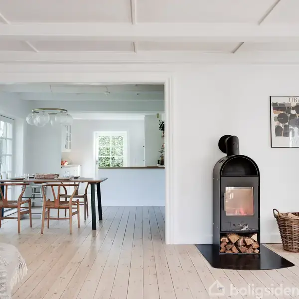 Brændeovn står varmende i et lyst, moderne stueområde med trægulv, åben ind til spisestue med træstole og bord. En kurv med brænde står ved siden af. En plakat hænger på væggen.