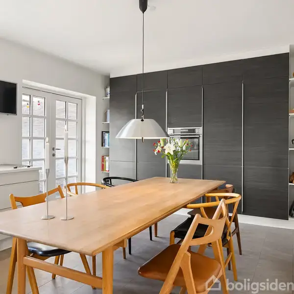Et spisebord med træstole står i køkkenet. Bordet har en vase med blomster. Baggrunden har hvide skabe, en ovn integreret i en mørk væg og en reol. En lampe hænger over bordet.