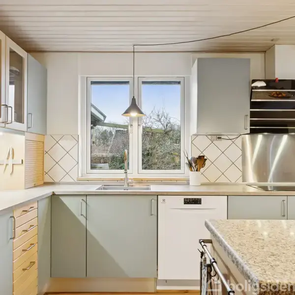 Et lyst køkken med grå skabe og skuffer, hvid opvaskemaskine, rustfri stål emhætte og køkkenvask under et vindue. Omgivet af hvide vægge og fliser.