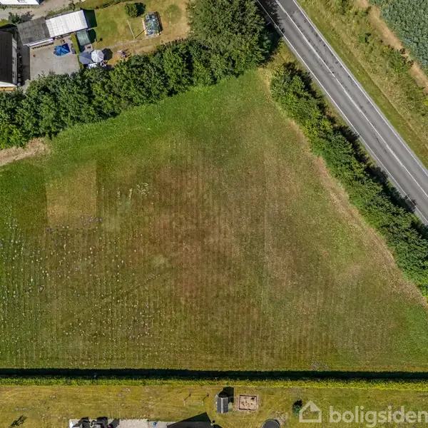 En trekantet græsplæne omgivet af hække og veje. Til venstre ses et hus med terrasse, mens en vej løber langs højre side. Grønne områder grænser op til plænen.