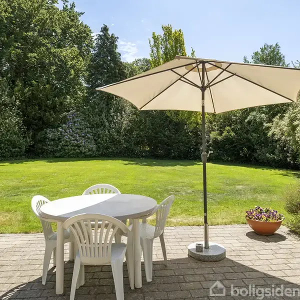 Rundt bord med fire hvide plastikstole står på en terrasse. En stor parasol giver skygge. Baggrunden viser en stor grøn græsplæne omgivet af buske og træer.