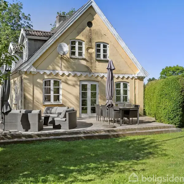 Et gult hus med saddeltag har en terrasse med havemøbler og parasoller. Terrassen er omgivet af en velplejet græsplæne og tætklippede hække, under en blå himmel.