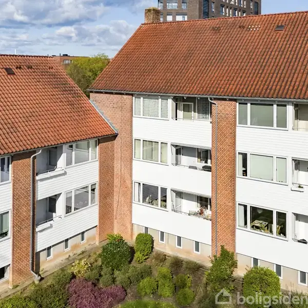 En rødtegltaget boligblok står stille i et grønt område med buske foran. Bygningen har flere etager med altaner, placeret under en lys blå himmel med spredte skyer.