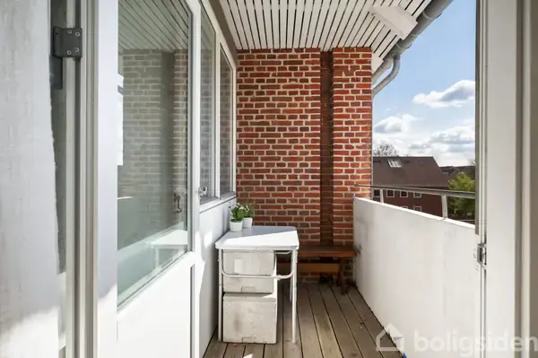 En lille altan med et hvidt bord og en potteplante står i hjørnet. Røde murstensvægge og en hvid gelænder omkranser rummet, mens solen skinner på vinduerne.