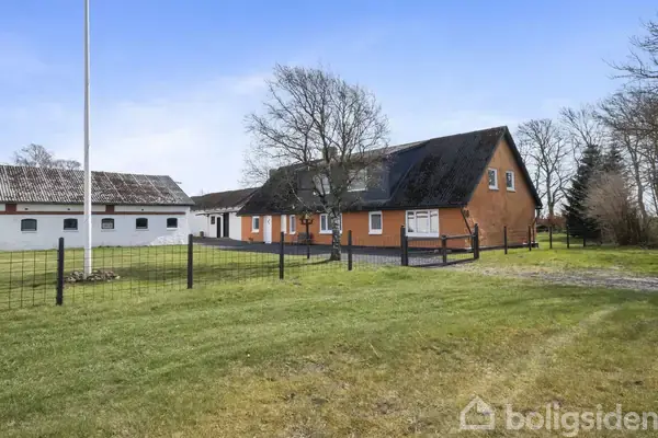 En rød murstensbondegård står stille omgivet af en grøn græsplæne og et hegn, med en tilstødende hvid bygning under en blå himmel.