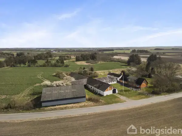 En gård med flere bygninger står langs en vej. Bygningerne er omgivet af marker og skove under en blå himmel.