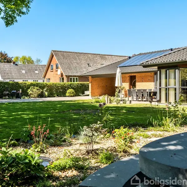 Hus med solpaneler på taget står i en velplejet have med græsplæne og blomsterbede. Terrassen har siddemøbler, omgivet af grønne træer og hække under en klar blå himmel.