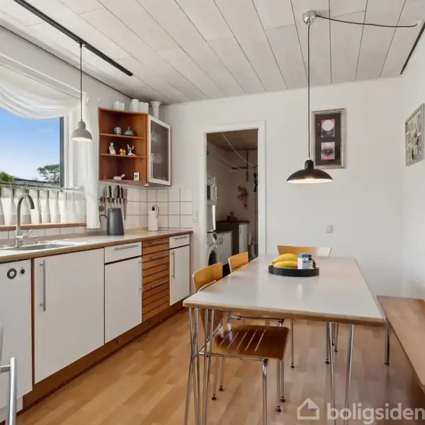 Et lyst køkken med træmøbler; en lang bordplade med vask og knivblok, et rektangulært spisebord omkranset af stole og bænk, hængende lamper. Store vinduer giver dagslys.