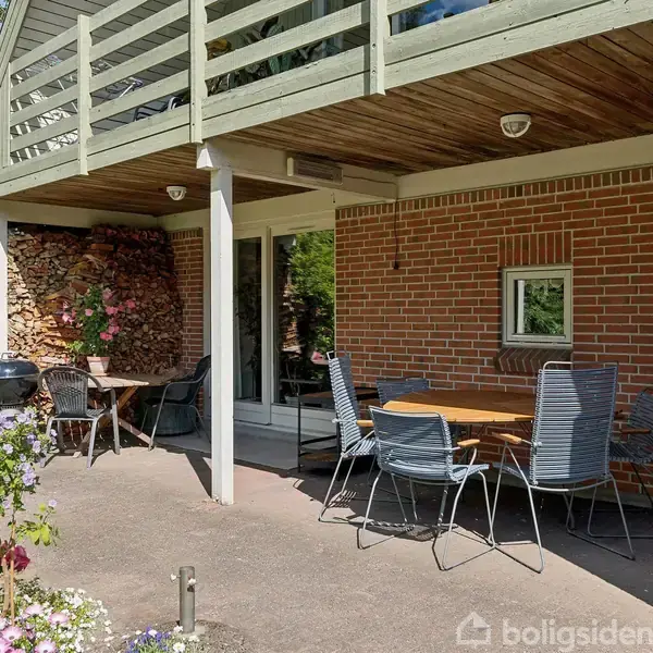 Terrassebord med stole står på en overdækket terrasse foran et murstenshus. Omkring bordet ses blomster og brændestabel. I baggrunden er der en grøn have med buske og træer.