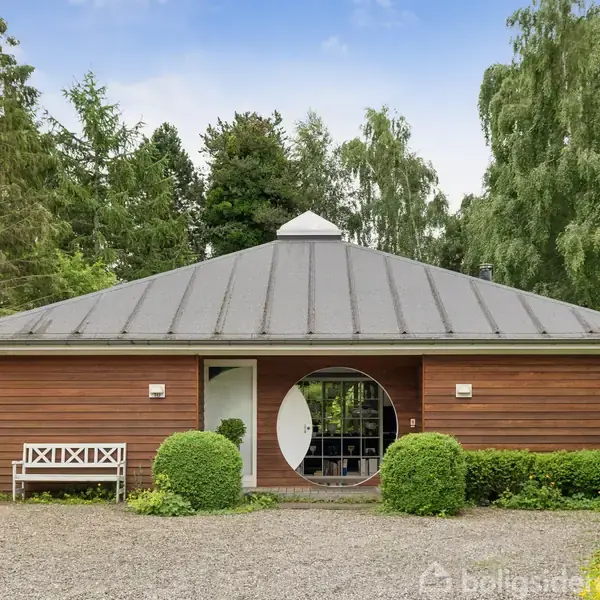 Et moderne hus med træbeklædning står omgivet af en have med buske og træer. Foran huset er der en hvid bænk ved siden af en grusbelagt indkørsel.