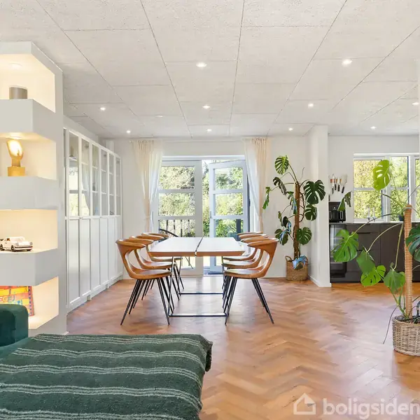 En moderne stue har et spisebord med seks stole. Store vinduer og grønne planter skaber en lys, indbydende atmosfære. En moderne grøn sofa og fladskærms-tv ses også.