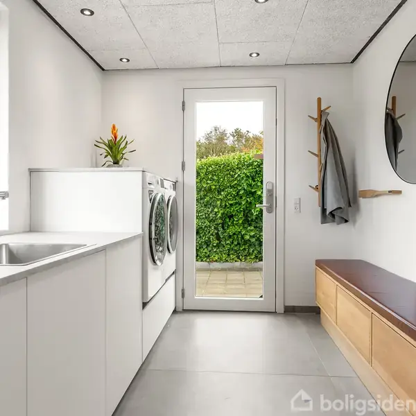 Vaskemaskine og tørretumbler står side om side i et lyst bryggers med hvide skabe. En bænk med opbevaring og et rundt spejl hænger ved siden af en jakke på knager.