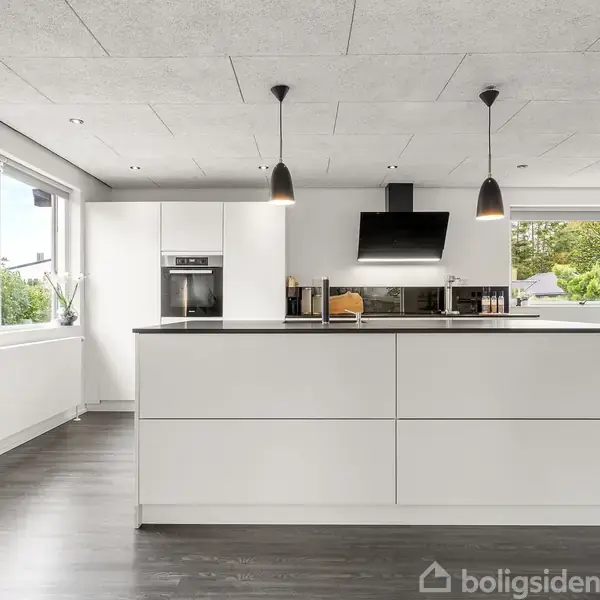 Et moderne, hvidt køkken med en stor ø, hængende lamper og integrerede hvidevarer. Vinduet til venstre viser udsigt til en have med grønne træer.