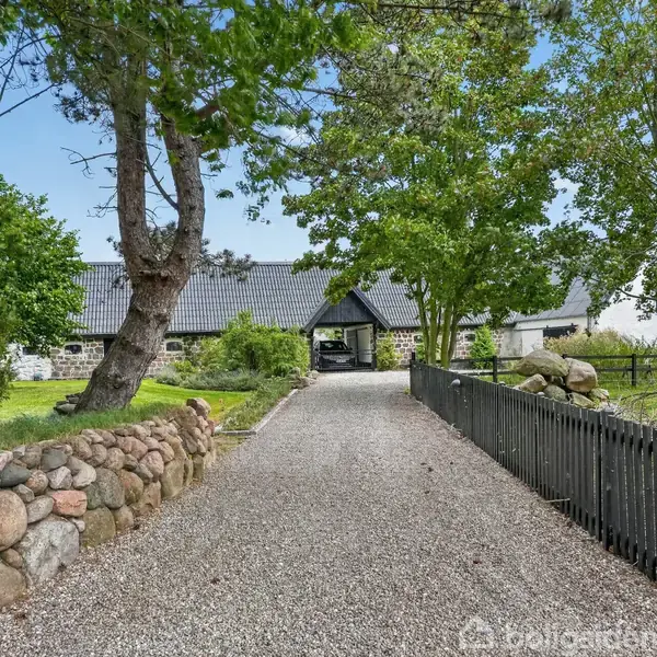 Gruset gårdsplads omgivet af hvide bygninger med sort tag. I midten dekoreret med planter og krukker. Blå himmel og grønne træer i baggrunden.