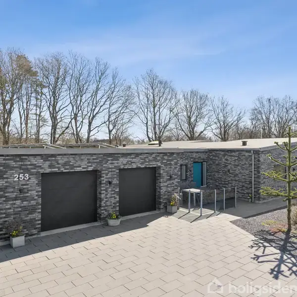 En moderne bygning står stille i et skovklædt område. Husnummer 253 er synligt. Bygningen har en række store, sorte garagedøre og en blå hoveddør.