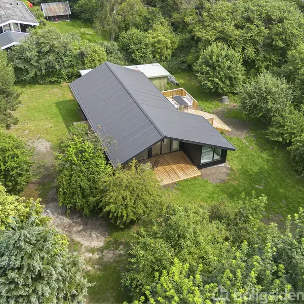 Et sort, moderne hus med tagterrasse omgivet af tæt, grøn vegetation. Huset har en stor terrasse, og der ses en grusvej ved siden af.