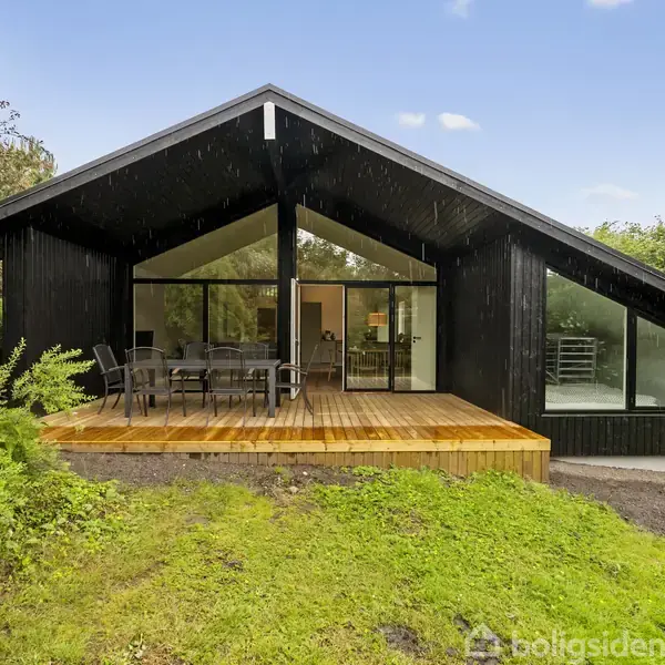Sort træhus står stille på en græsplæne omgivet af grønne buske og træer. En åben terrasse med havemøbler strækker sig fra huset under en blå himmel.
