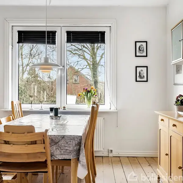 Et lyst spisestuemiljø med et træbord dækket med en dug og omgivet af træstole. En lampe hænger over bordet. Vindue med udsigt til en have. Malerier hænger på væggen.