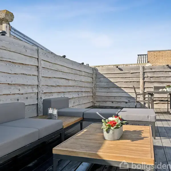 Grå udendørs sofaer står langs grå trævægge. Et lavt træbord med en blomsterdekoration står imellem dem. Bagved ses et højere bord med stole på en træterrasse.