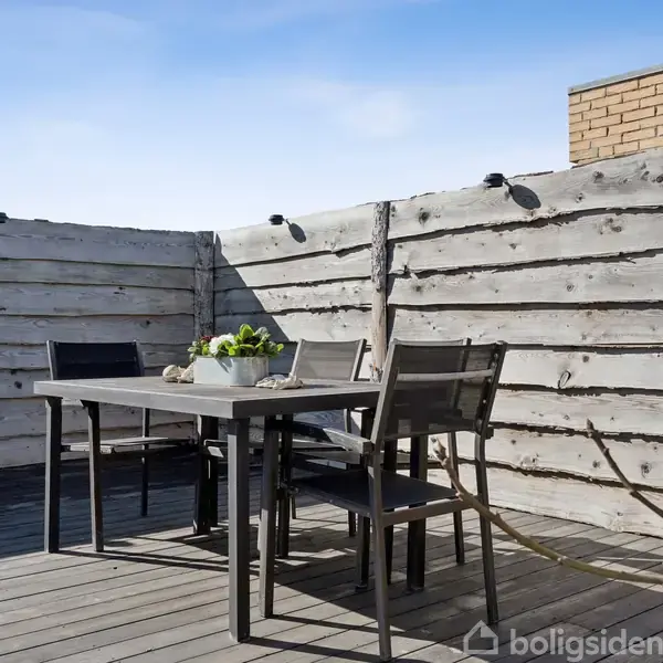 Et bord med fire stole står på en terrasse med grå træplanker. Bordet har en plante som dekoration. Baggrunden består af en høj, lys træhegn i et udendørs miljø.