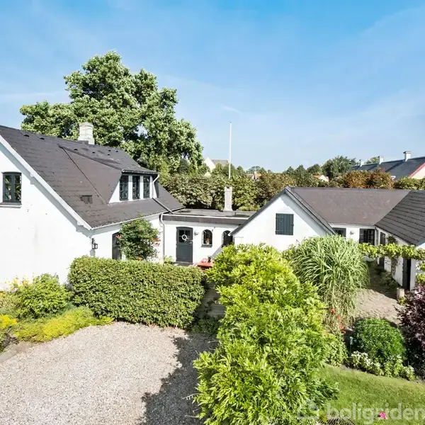 Hus med hvid facade og sort tag står i velplejet have med buske og træer. Baggrunden viser klare blå himmel og store grønne træer.