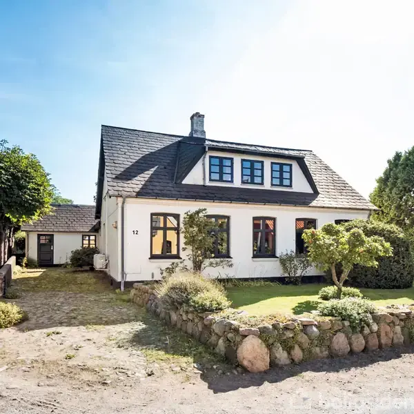 Et hvidt hus med mørkt tag står i en have med en stenmur, omgivet af træer og buske. "12" ses på husets væg. En grussti løber langs grunden.