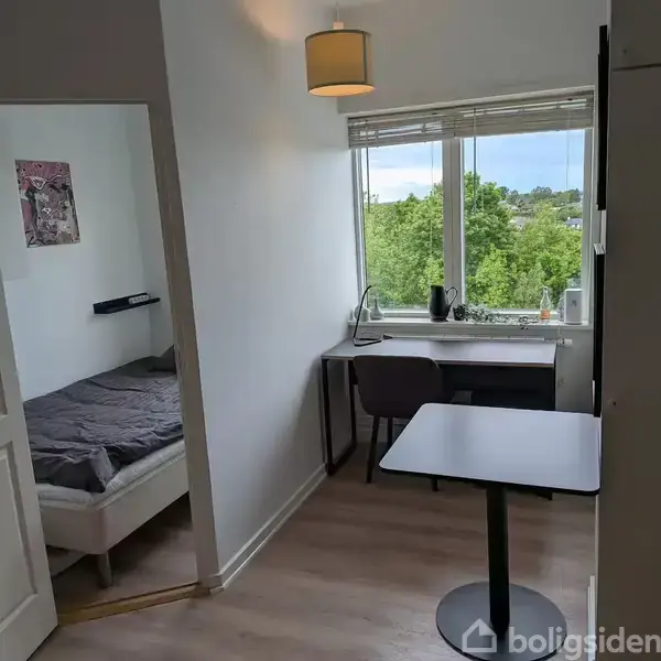 Stue med skrivebord og stol foran et vindue med udsigt til træer. På højre side ses et lille bord. Et åbent soveværelse med seng er til venstre.