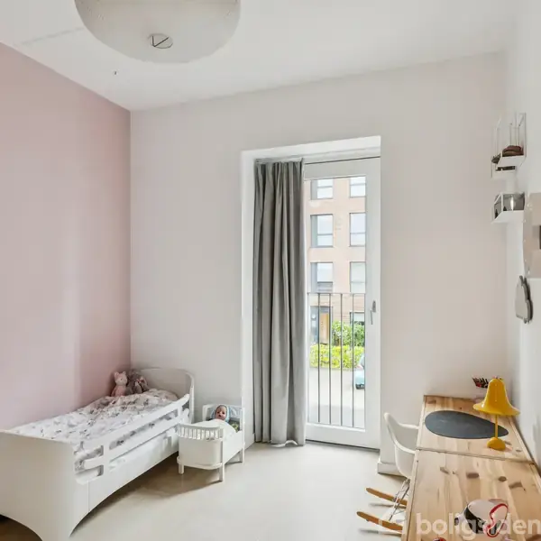 Børneværelse med en hvid seng til venstre og et lille skrivebord med stol til højre. En stor glasdør med grå gardiner fører ud til en balkon.