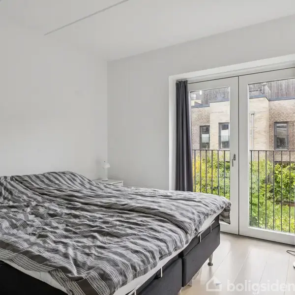 Soveværelse med dobbeltseng med stribet sengetøj; ved siden af vinduer åbne mod en balkon med udsigt til bygninger og grønne træer. Hvide vægge og en gulvlampe ved balkon.