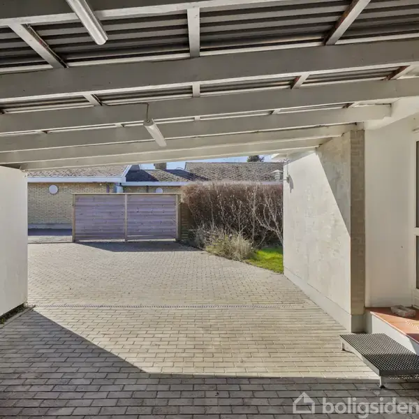 En carport med et fladt tag og lyse vægge er tilsluttet et hus. I baggrunden ses en brolagt indkørsel og en lukket træport.