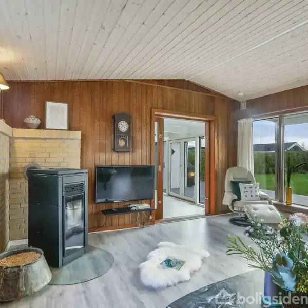 En stue med en sort ovn og en vægmonteret fladskærm ved pejsen. Store vinduer giver udsigt til en have med terrasse.