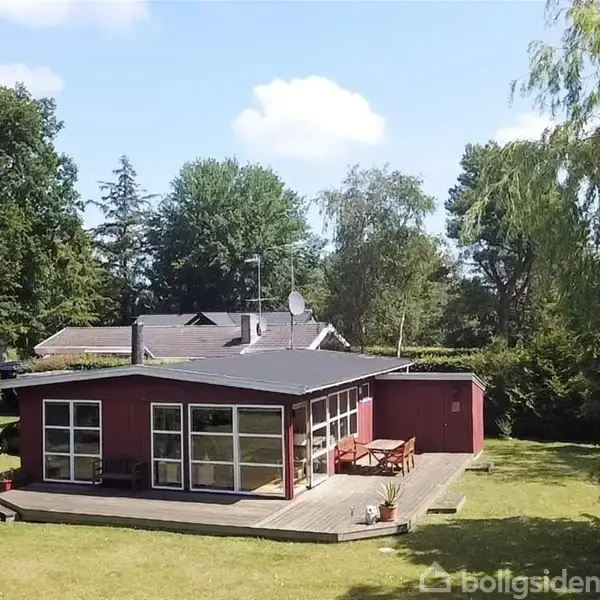 Et rødt træhus står på en stor, grøn græsplæne omgivet af træer. Foran huset er der en terrasse med et bord og stole.
