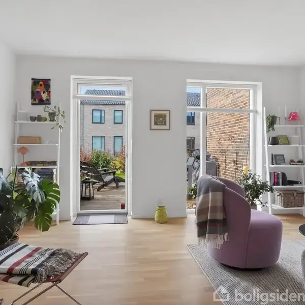 En lys stue med åbne døre til en altan. Stuen har en lilla lænestol, grønne planter, fladskærms-tv og hylder med bøger. Udenfor ses en terrasse med møbler.