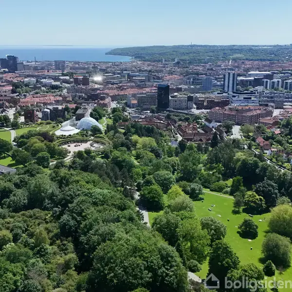 En storby med parker og grønne områder, herunder et drivhus, udfolder sig under en klar blå himmel. Bygninger og havet strækker sig i baggrunden, og viser en blanding af natur og urbanitet.