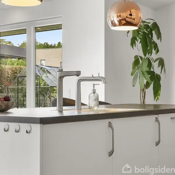 Køkkenø med hvid bordplade står centralt; en skål med frugter og en vandmelon placeres ovenpå. En grøn plante ved siden. Store vinduer viser en udendørs have med drivhus.