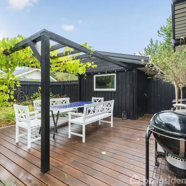 Terrasse med havemøbler under en pergola dækket af planter. En grill står til højre. Omkring er der sorte trævægge og grøn vegetation, skaber en hyggelig udendørs atmosfære.