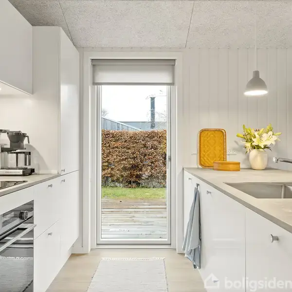 Et lyst køkken viser en kaffemaskine placeret på en hvid bordplade. En dør åbner ud til en terrasse. Blomster i en vase står ved siden af vasken.