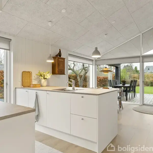 Køkken med hvid køkkenø, vase med blomster og håndvask, omgivet af store vinduer, der giver udsigt til en grøn have. Spisebord ses i baggrunden.