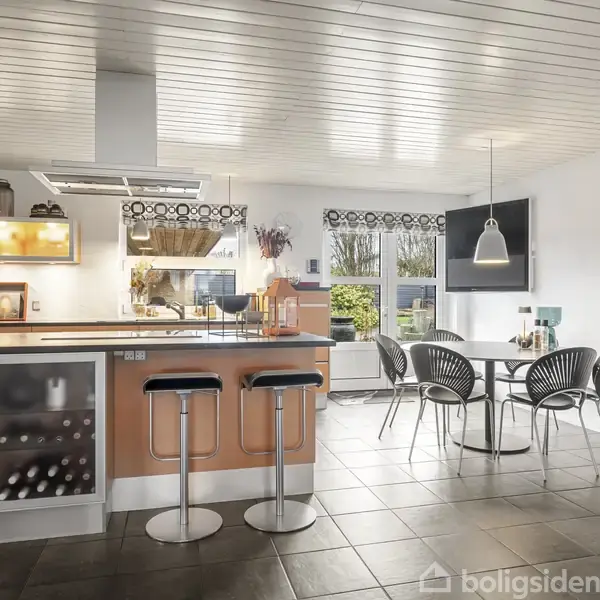 Køkken med barstole omkring en ø, fyldt med køkkenredskaber og vinflasker. I baggrunden ses et spisebord med stole ved siden af vinduer, der lukker naturligt lys ind.