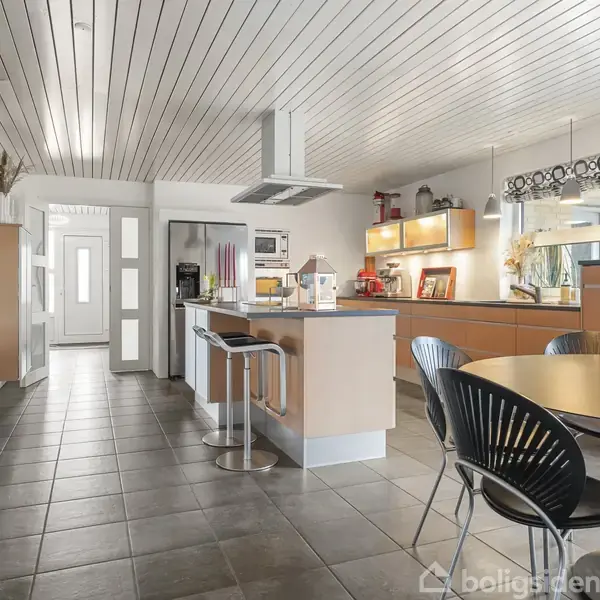 Et moderne køkken med lyse træskabe, mørke flisegulve og et rundt spisebord med sorte stole. Rummet er lyst med hængende lamper og vindue. En bar med høje stole ses midt i køkkenet.