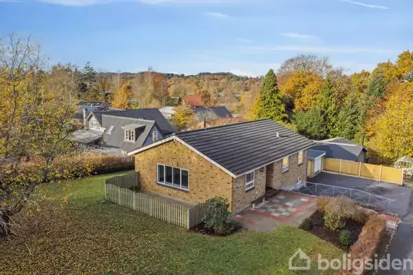 Et enkeltplans murstenshus står i en velplejet have med græsplæne og træer i efterårsfarver. En indkørsel og et hegn omgiver huset i et roligt nabolag.