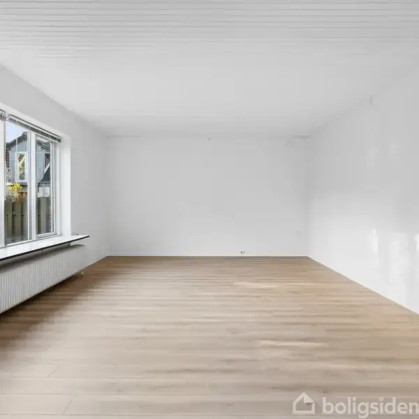 Et tomt rum med trægulv har et stort vindue til venstre, der slipper naturligt lys ind. Hvide vægge og en radiator under vinduet. Dør til højre.