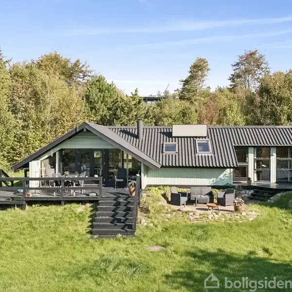 Et sommerhus i træ med sort tag og store vinduer står i en grøn have. Huset har en terrasse med trapper i forgrunden, omgivet af træer.