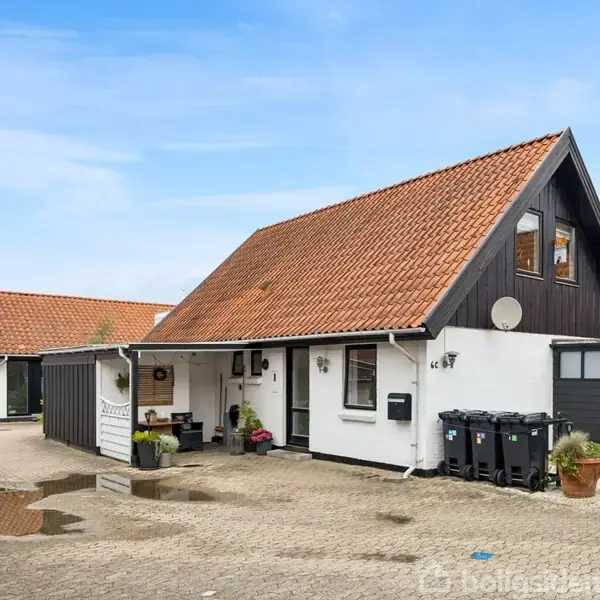 Hus med rødt tegltag og sort træbeklædning, beliggende på brostensbelagt gårdsplads med planter og affaldscontainere. Skyfri himmel og naboer i baggrund. Adresse: 16 C.