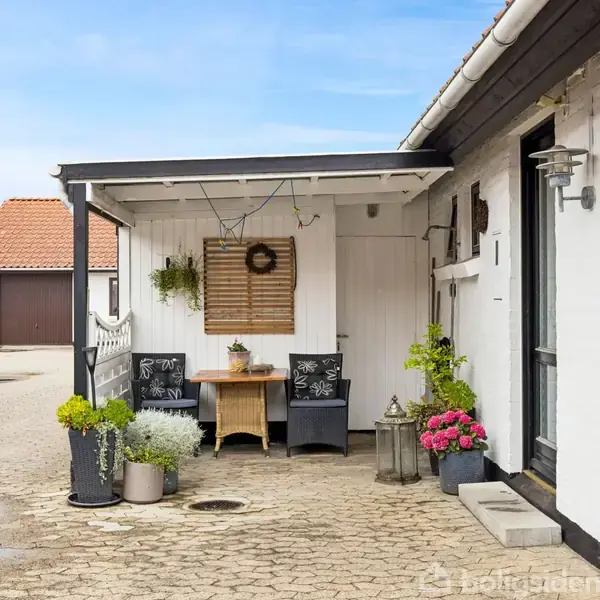 Overdækket terrasseområde med bord og to stole; omgivet af hvide bygninger med røde tage. Blomster i krukker og en væghængt dekoration giver et hyggeligt udseende.