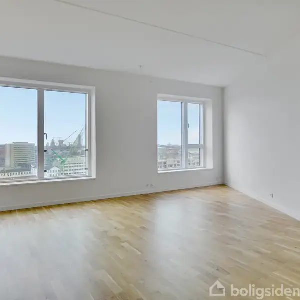 Tomt rum med lyst trægulv, to store vinduer, der giver udsigt til byens skyline. Væggene er hvide, og rummet er minimalistisk og uindrettet, hvilket skaber en lys og åben atmosfære.