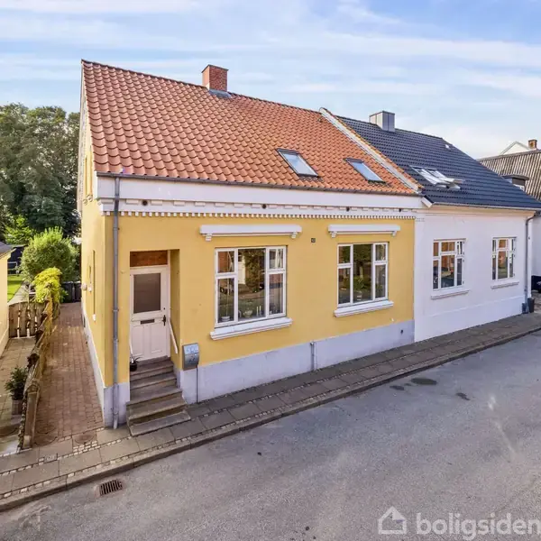 Gult hus med rødt tegltag står i en bygade, omgivet af lignende huse. To sorte biler er parkeret på den asfalterede vej ved siden af huset.