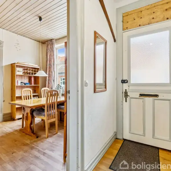 En entrédør åbner til en lille gang med trægulv, hvorfra en spisestue med træbord og stole er synlig. Rummet er lyst med et vindue og loftslampe.