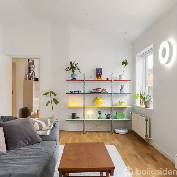 En stue med en grå sofa og farverige hylder fyldt med planter og dekorationer. En træbord står foran sofaen, og store vinduer giver naturligt lys i rummet.
