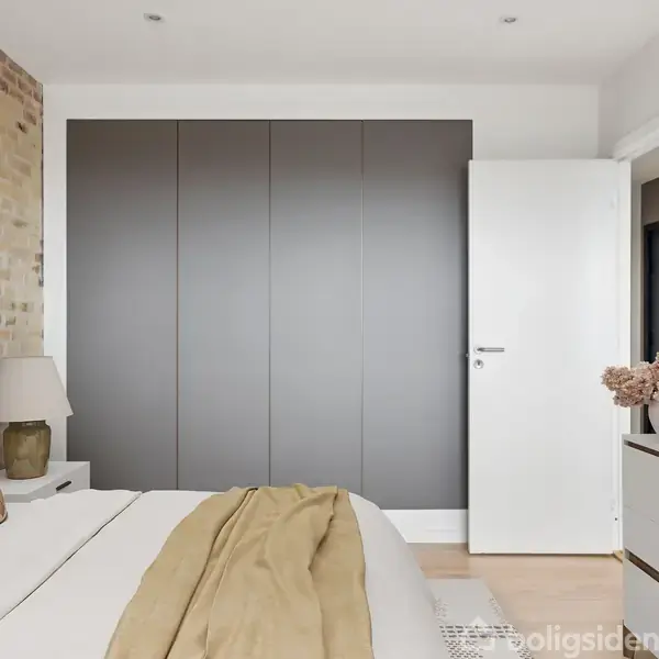 Soveværelse med seng dækket af beige sengetøj, ved siden af natbord med lampe. Skydedørsskab og åben dør i baggrunden. Indretningen er minimalistisk med en murstensvæg.